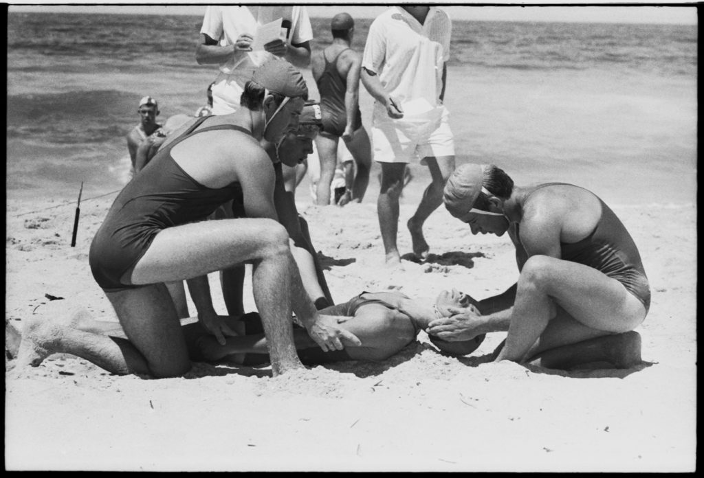 Black and white photograph showing three lifesavers wearing full strapped costumes and caps on the water's edge kneeling over a man on whom they are demonstrating resuscitation.